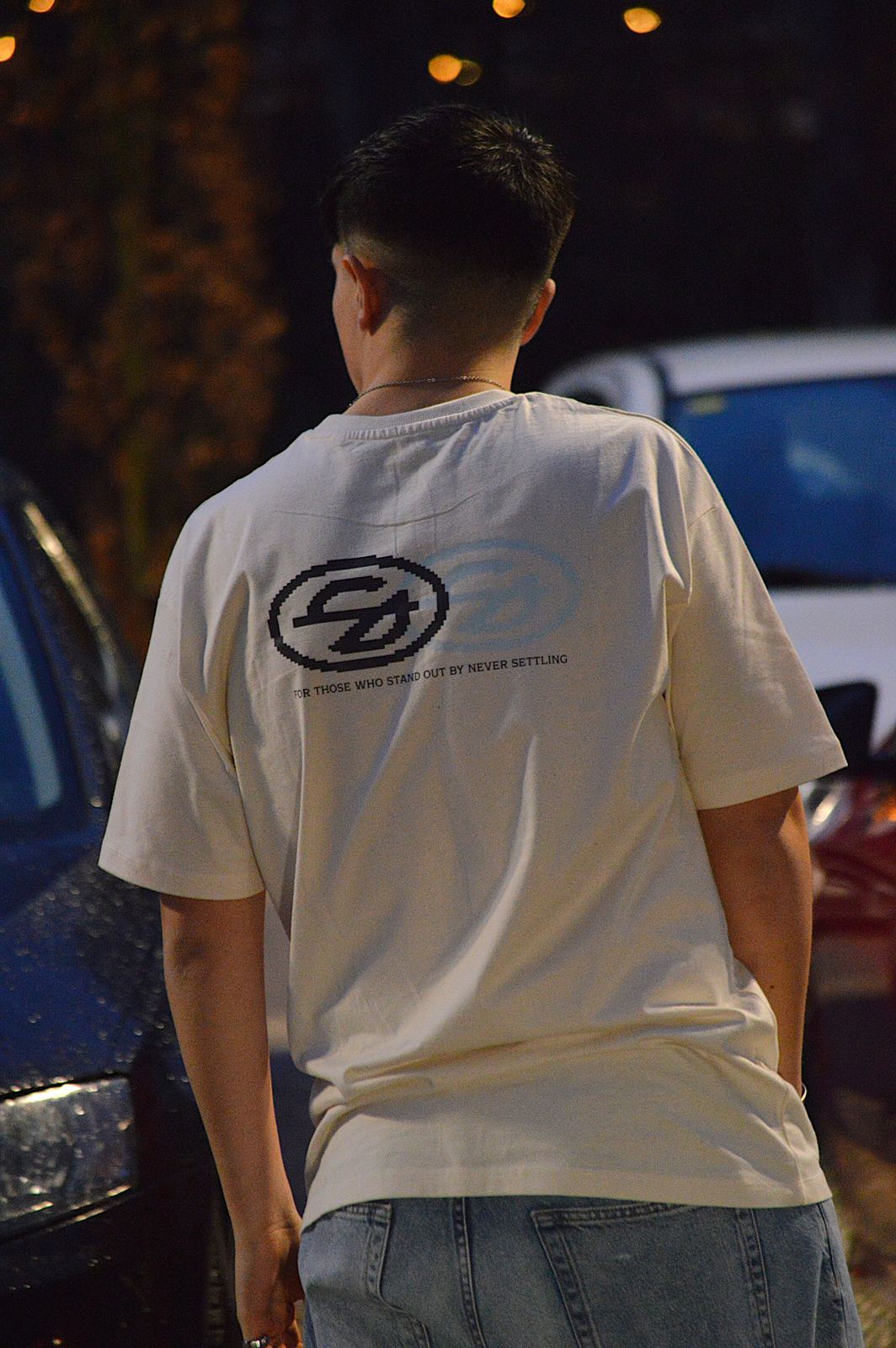 Modelo de espaldas con la camiseta Logo color crema y la frase For those who stand out by never settling